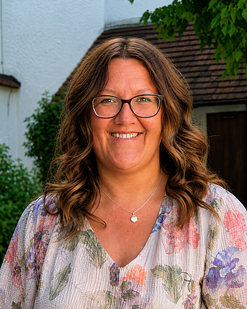 Porträt einer lächelnden Frau mit Brille, braunen lockigen Haaren und floralem Oberteil vor Hausfassade und Grün. Erste Jugendbeauftragte der Stadt Monheim: Kathrin Böswald