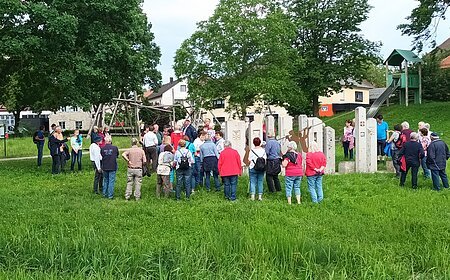 Gruppe von Menschen steht im Kreis auf einer Wiese, umgeben von Bäumen und Gebäuden. Im Hintergrund eine große Rutsche zu sehen.