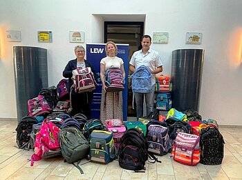 Drei Personen stehen hinter einer großen Anzahl verschiedener Schulranzen in einem hellen Raum. Die Übergabe der gesammelten Schulranzen in Monheim zur Aktion vom LEW "Ranzen her".