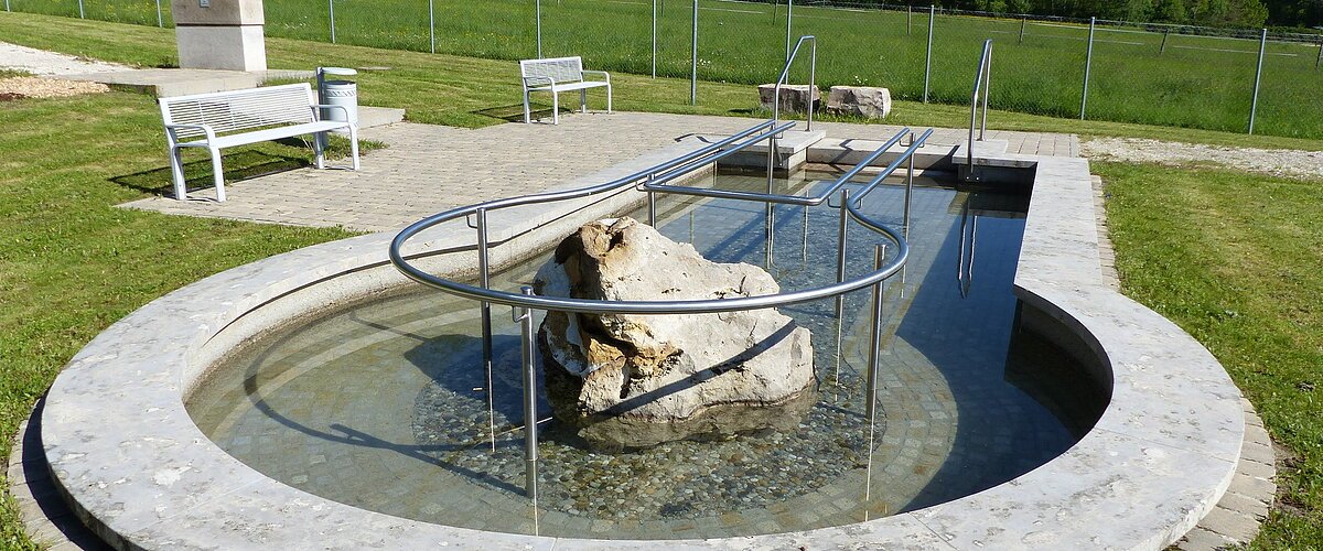 Rundes Kneippbecken in Monheim mit einem Geländer und einem Barfußpfad, in der Mitte ein großer weißer Stein. Umgeben von einer Wiese, darauf Sitzmöglichkeiten und eine große Grünfläche mit Bäumen hinterhalb, abgetrennt mit einem Zaun.