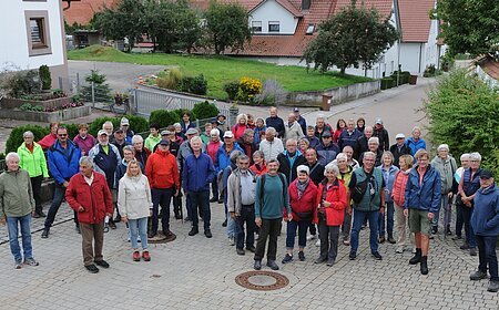Gruppe von etwa 50 Personen steht auf gepflastertem Platz in einem Wohngebiet mit Häusern und Bäumen im Hintergrund. Senioren wandern mit SoMit. Nächste Wanderung am 14.10.2026 in Huisheim/Gosheim