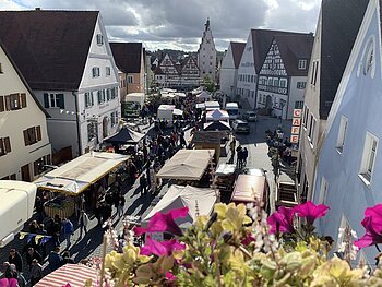 Blick von oben auf belebten Markt mit Ständen und Menschen in einer Altstadt mit Fachwerkhäusern und Café.