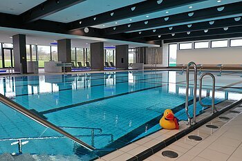 Innenansicht eines Hallenbades mit leeren Liegestühlen, einem befüllten Schwimmbecken und einer großen gelben Gummiente am Beckenrand. An der Decke Leucht-Spots und im Hintergrund große Fenster.