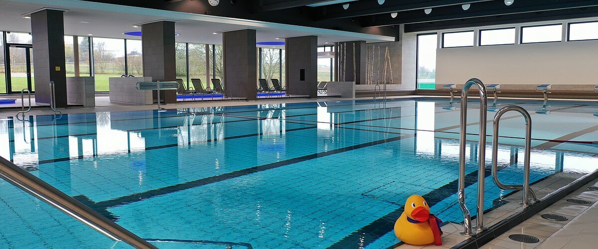 Innenansicht eines Hallenbades mit leeren Liegestühlen, einem befüllten Schwimmbecken und einer großen gelben Gummiente am Beckenrand. An der Decke Leucht-Spots und im Hintergrund große Fenster.