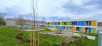 Zwei Gebäude des städtischen Kindergartens Monheim, eins davon mit einer bunten Fassade, davor ein Garten mit Spielgeräten Bäumen und Rasenfläche, bewölkter Himmel.