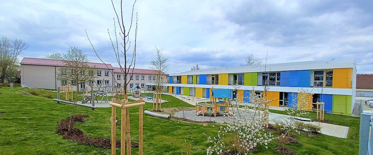 Zwei Gebäude des städtischen Kindergartens Monheim, eins davon mit einer bunten Fassade, davor ein Garten mit Spielgeräten Bäumen und Rasenfläche, bewölkter Himmel.